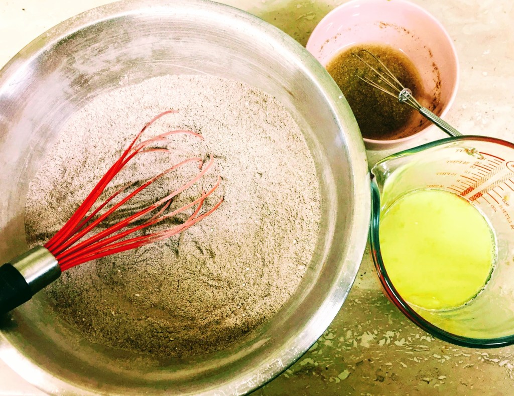 The ingredients before they are combined. The dry ingredients have been whisked together in a large mixing bowl, the flax seeds and water  have been mixed and left to sit in a smaller bowl, and the soya milk has been left to curdle in a mixing jug.