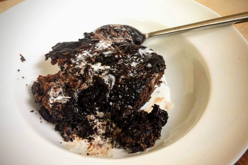 A portion of vegan and gluten-free saucy chocolate pud. The sponge is moist and laced with dark chocolate sauce, and smothered in vegan cream.