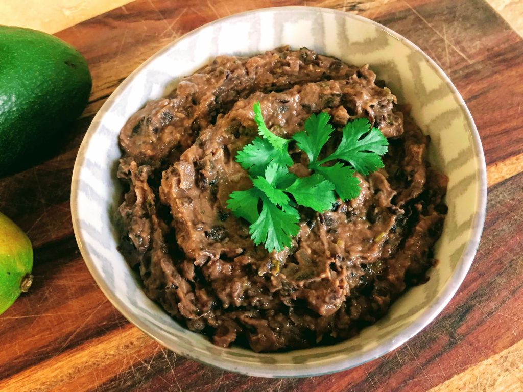 A bowl of creamy drunken re-fried black beans with a coriander leave garnish.