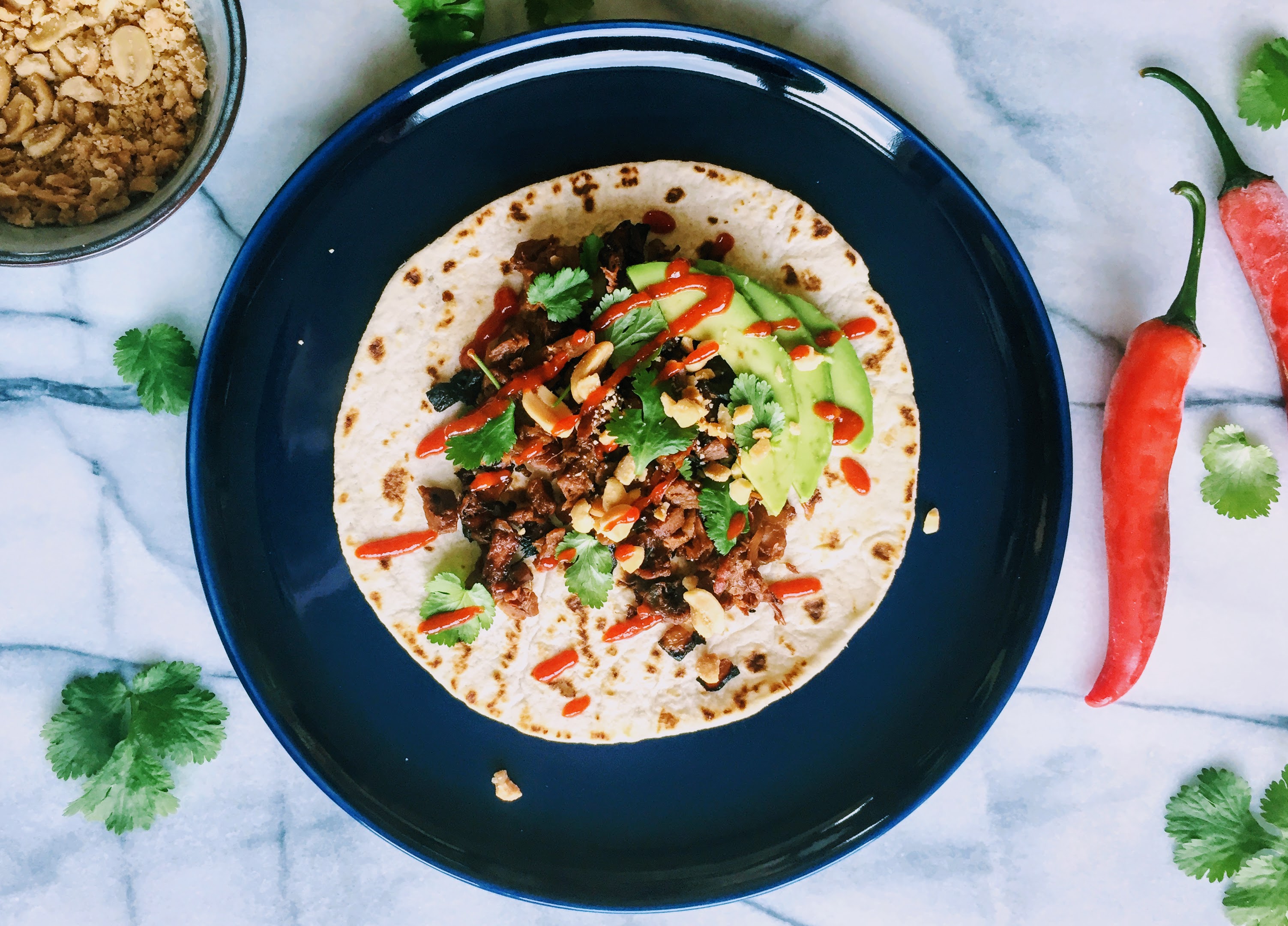 An aerial view of the jackfruit wrap; a tortilla wrap on a dark blue plate, piled with the tamarind pulled jackfruit, avocado slices, coriander leaves, crushed peanuts and drizzled in hot sauce. A small bowl of crushed peanuts and some red chillies are either side of the plate, and there are a few more coriander leaves scattered over the marble work surface.
