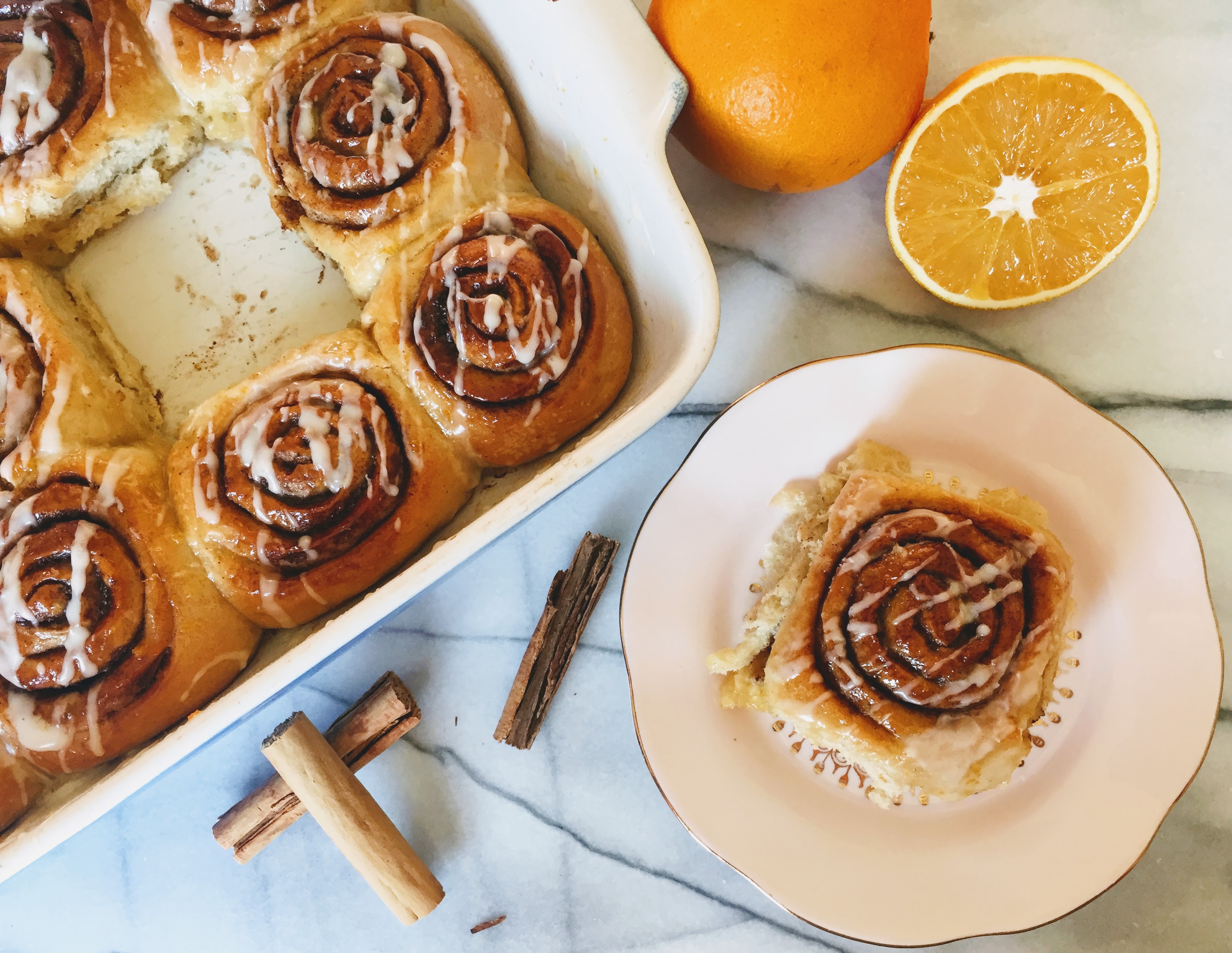 A cinnamon roll on a light pink plate, with a cinnamon swirl and a drizzle of white icing. The plate is next to the dish the with remaining cinnamon rolls, and there are cinnamon sticks scattered on the side and a bright cut orange.