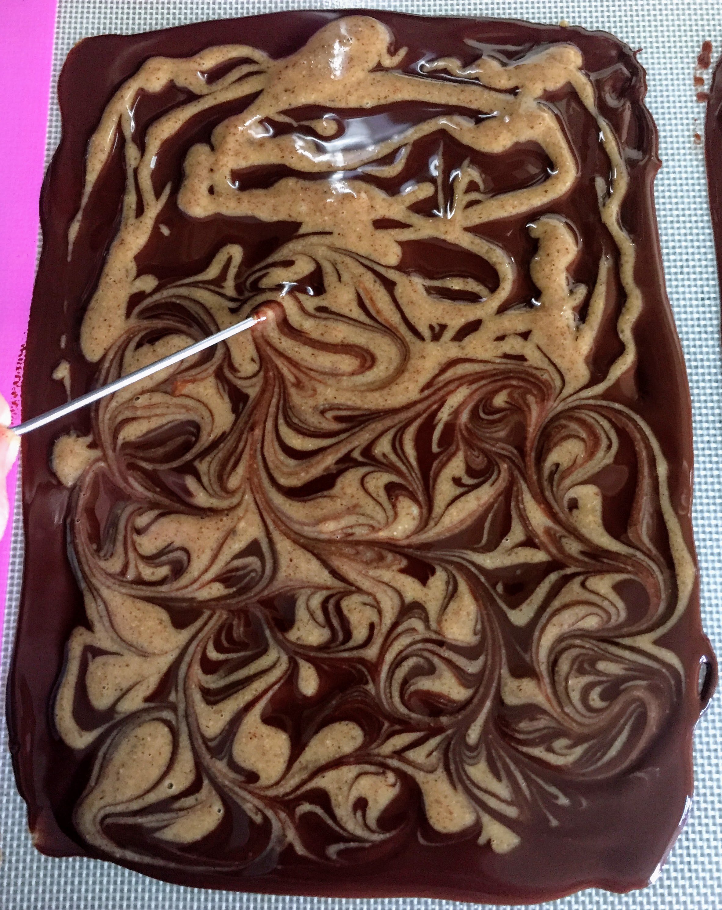 A rectangle of melted dark chocolate, spread on a baking tray and drizzled with almond butter mixture. The top half shows the almond butter roughly drizzled, and the bottom half shows the marbled effect when a skewer is run through the chocolate and almond mixture.