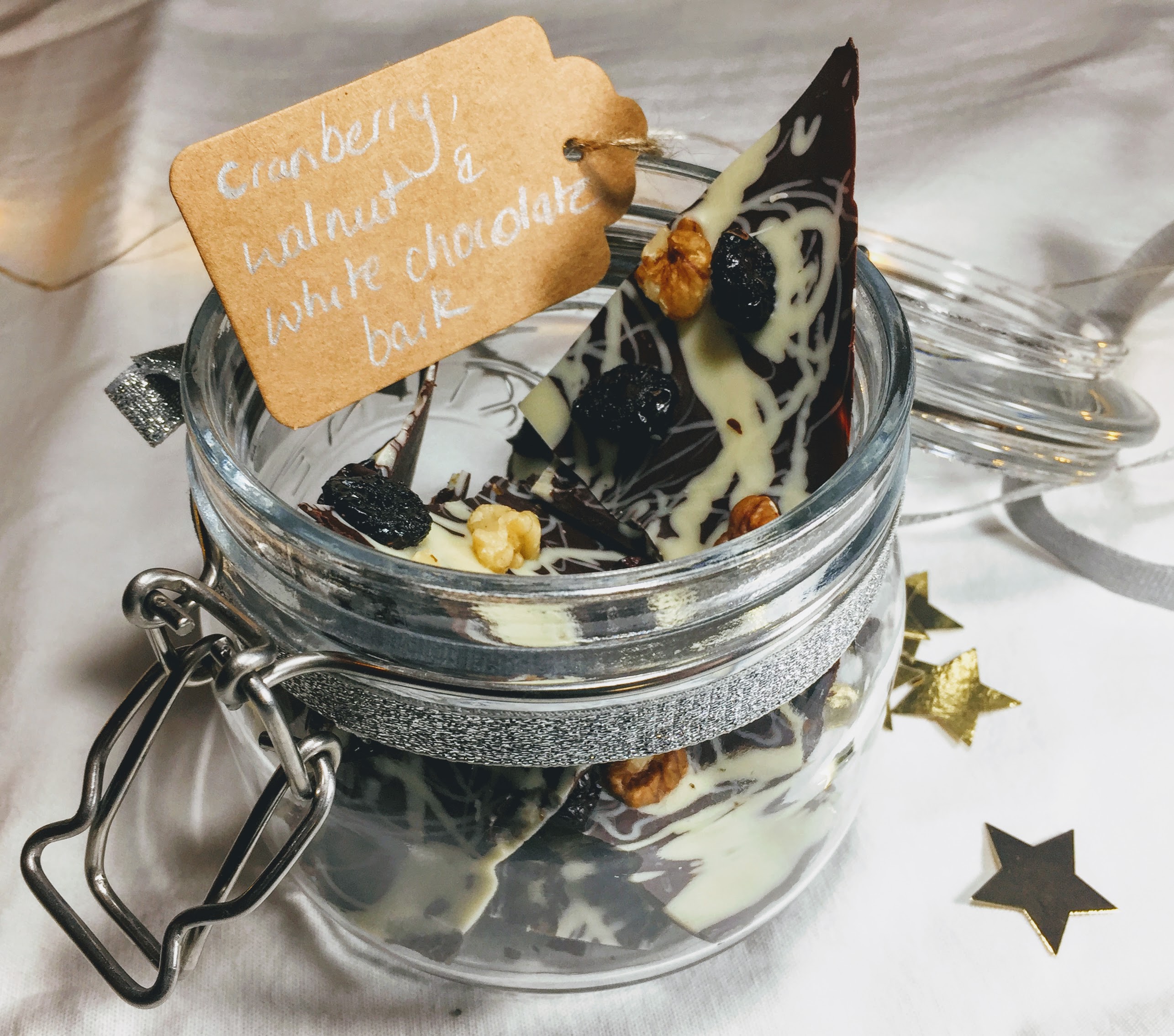 A small glass kilner jar filled with shards of chocolate bark. There jar has a glittery silver ribbon and a brown paper tag that reads 'Cranberry, walnut & white chocolate bark' handwritten in silver pen. 