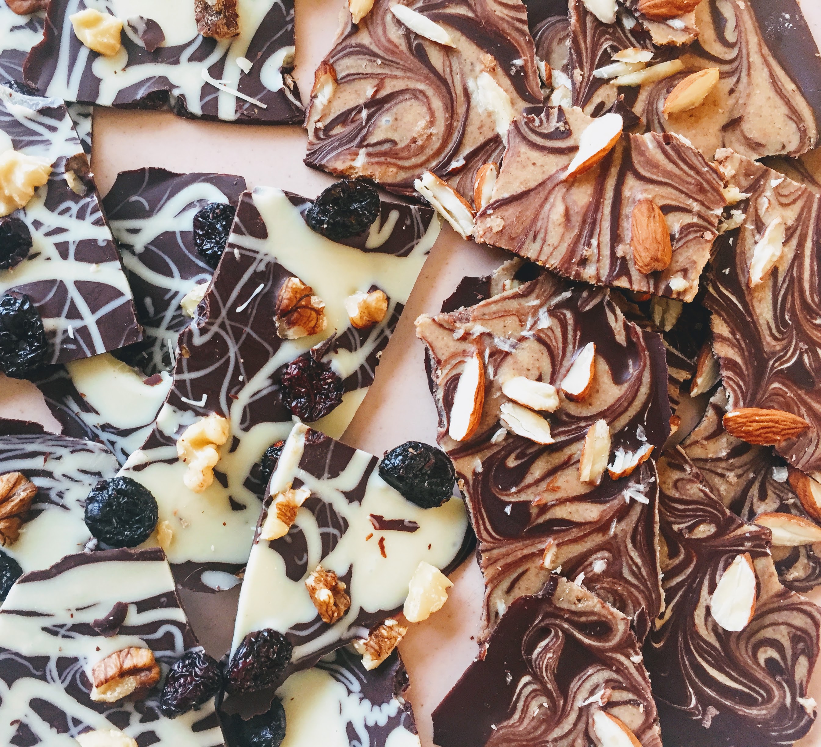 Shards of chocolate bark. On the left-hand side are thin pieces of dark chocolate melded with white chocolate and covered in dried cranberries and chopped walnuts. On the right hand side are thin pieces of dark chocolate marbled with almond butter, with slivered almonds. The pieces are broken and irregular and arranged on a light pink background. 