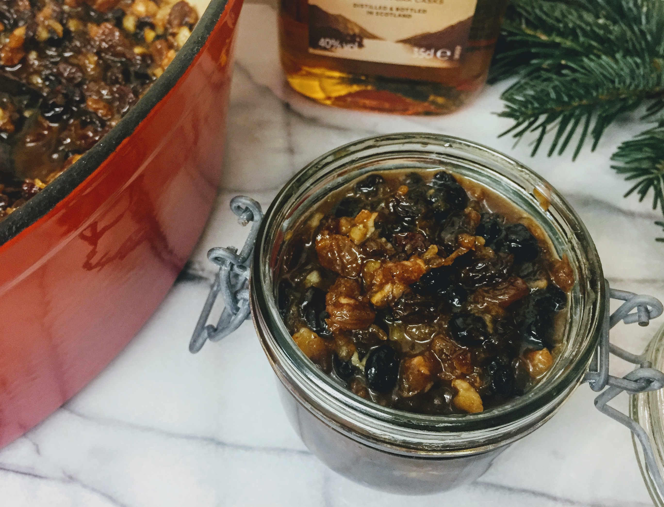 A small glass jar packed with mincemeat; the dried fruit is slightly plump from the liquid and is dark and glistening. The pan with the remaining mincemeat, a half bottle of whiskey and a pine sprig surround the jar.