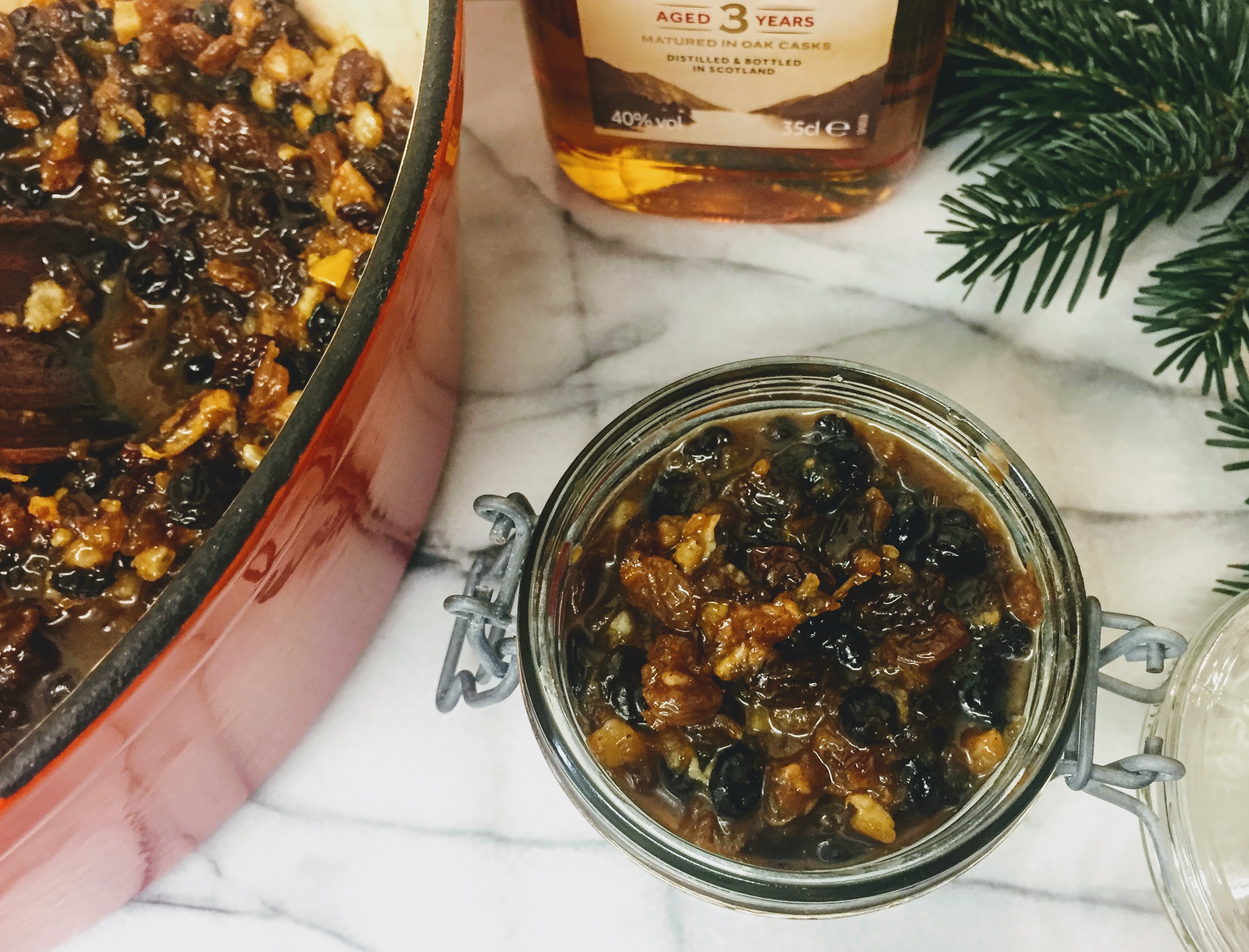 A birds-eye view of a small glass jar packed with orange and whiskey mincemeat. On the left there is a pan full of the remaining mincemeat, and at the top of the picture is a small bottle of aged scotch whiskey and  a sprig from a pine tree.