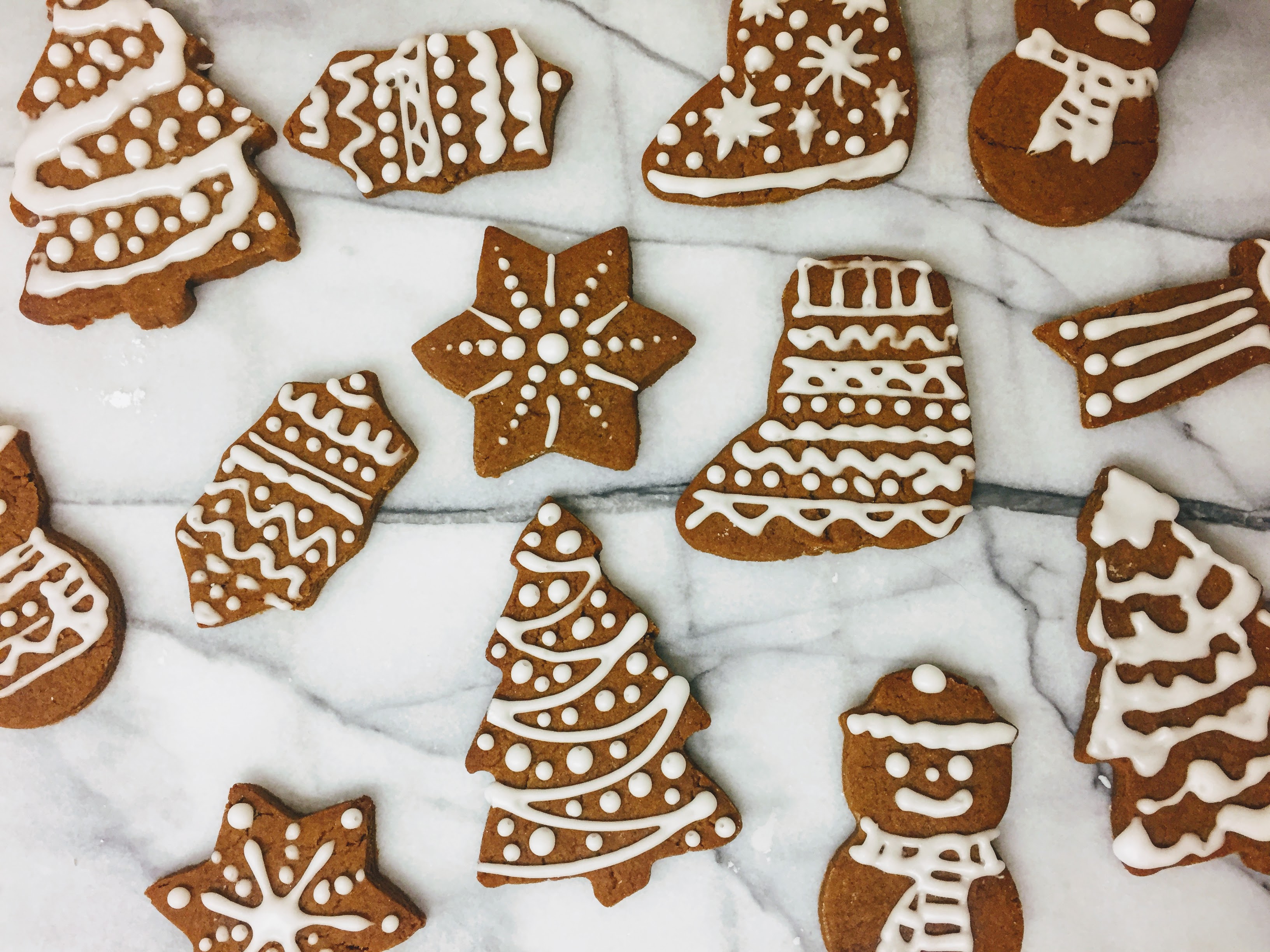 An aerial view of the gingerbread biscuits laid out on a marble board, shaped like stars, christmas trees, stockings, snowmen and holly. The biscuits are dark golden brown and are decorated with piped bright white icing. 