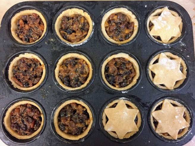 A cupcake tray - each hole is lined with pastry and filled with mincemeat, up to the rim. A few have their star-shaped lids pressed on.