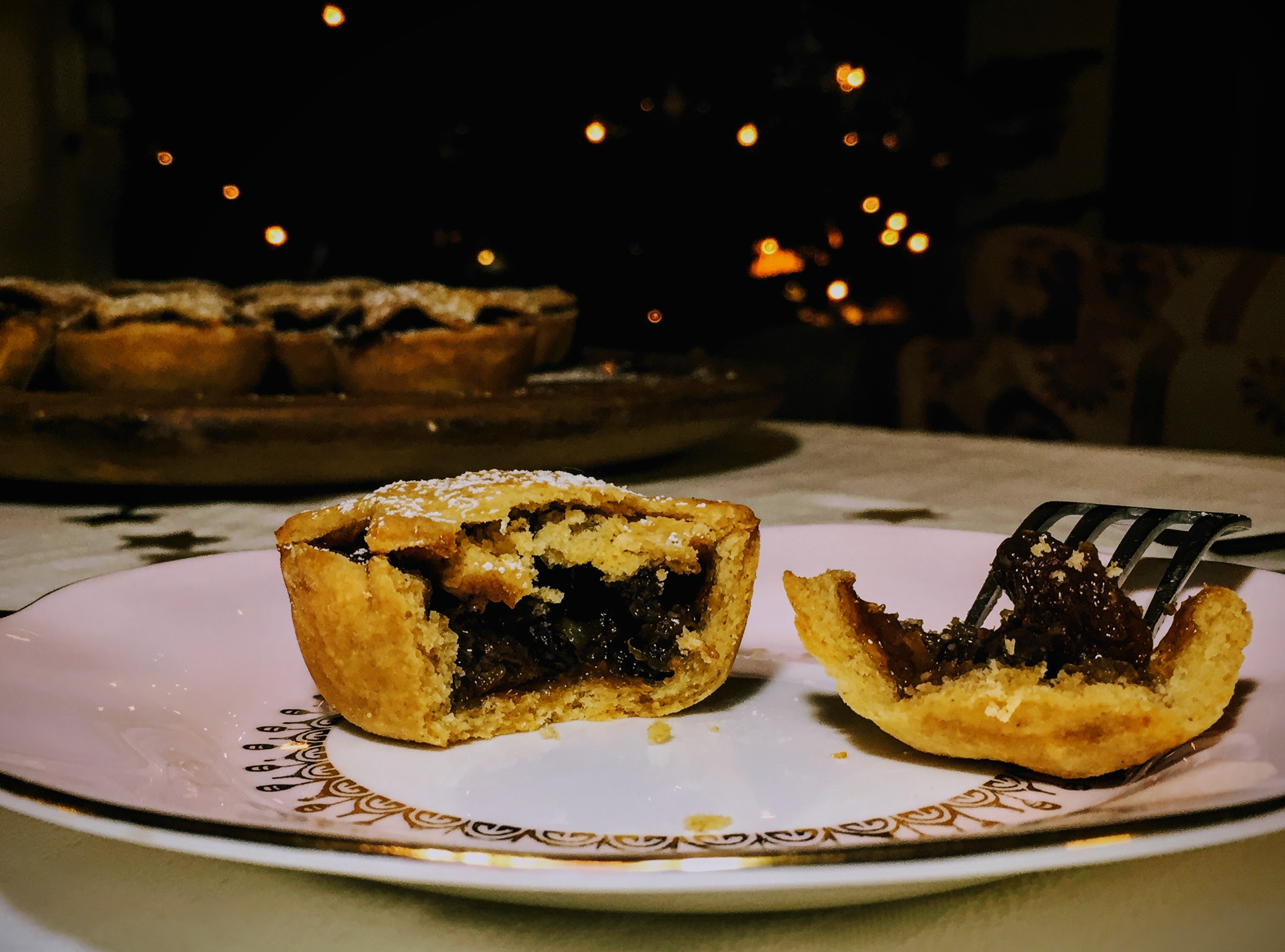A mice pie, cut open to reveal the dark and glistening mincemeat filling. The pastry is golden, flakey and crumbling. The rest of the mince pies are on a board behind, and Christmas lights sparkle in the background.