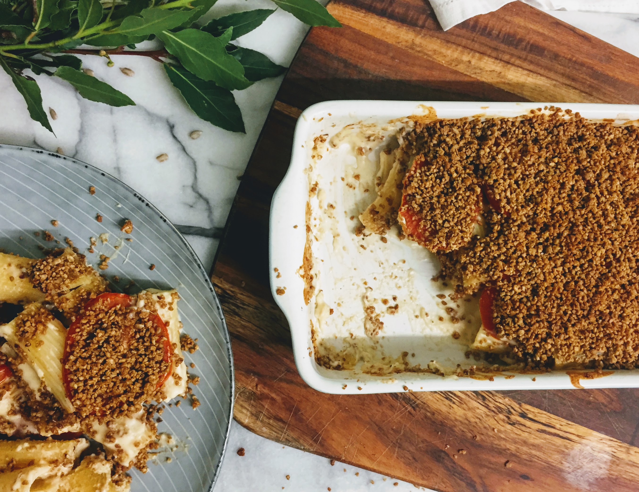 On the left is a pile of vegan macaroni cheese with a creamy white sauce and golden crumble topping, on a blue plate. On the right, an oven dish sits on a wooden board, and is filled with the baked cheesy pasta, with a few scoops removed.