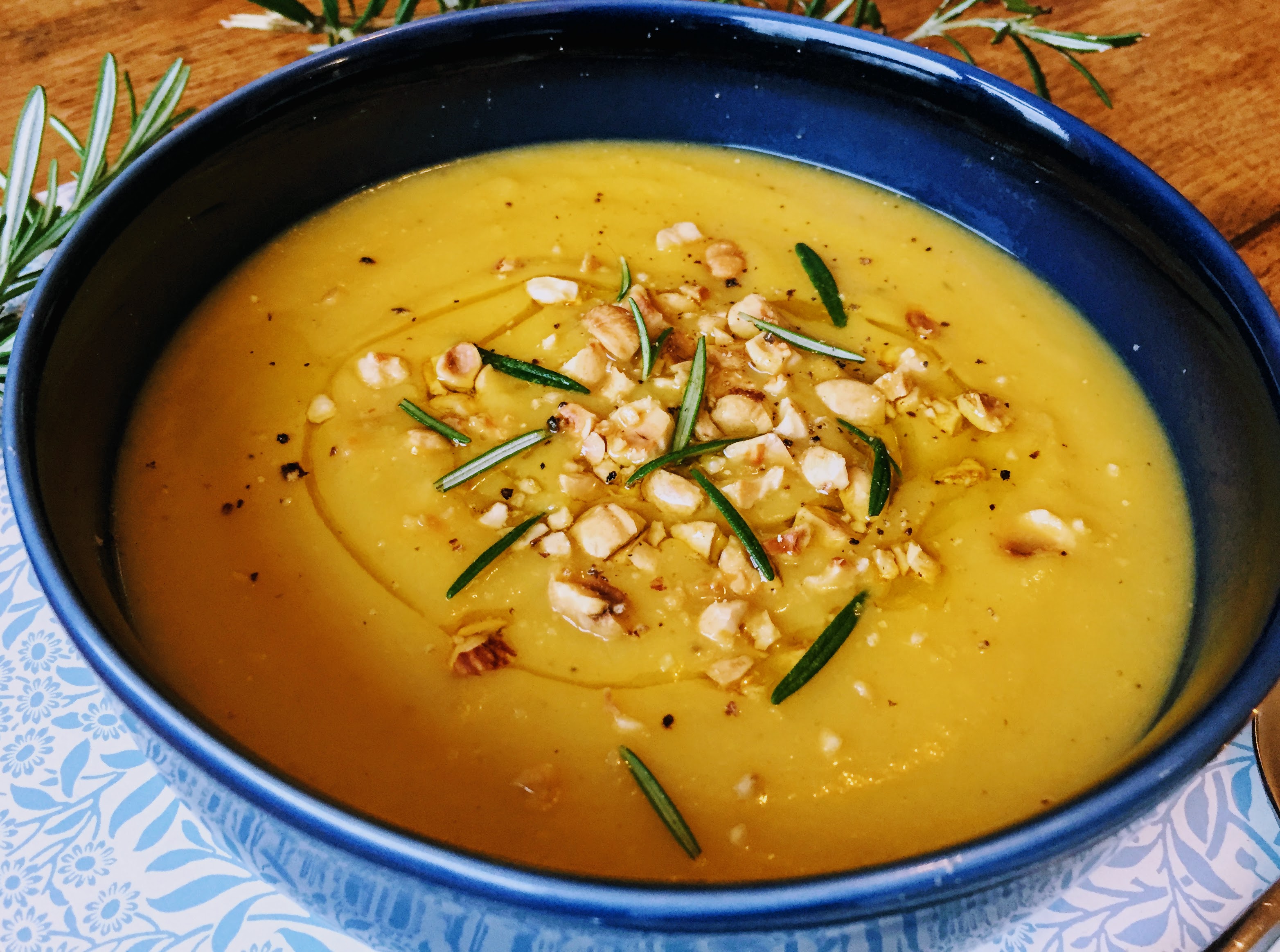 A close-up of the soup. The bowl is a dark blue which contrasts with the sunny orange of the soup. The soup is smooth and velvety, and you can see the swirls of drizzled olive oil beneath the floating crushed and toasted hazelnuts.