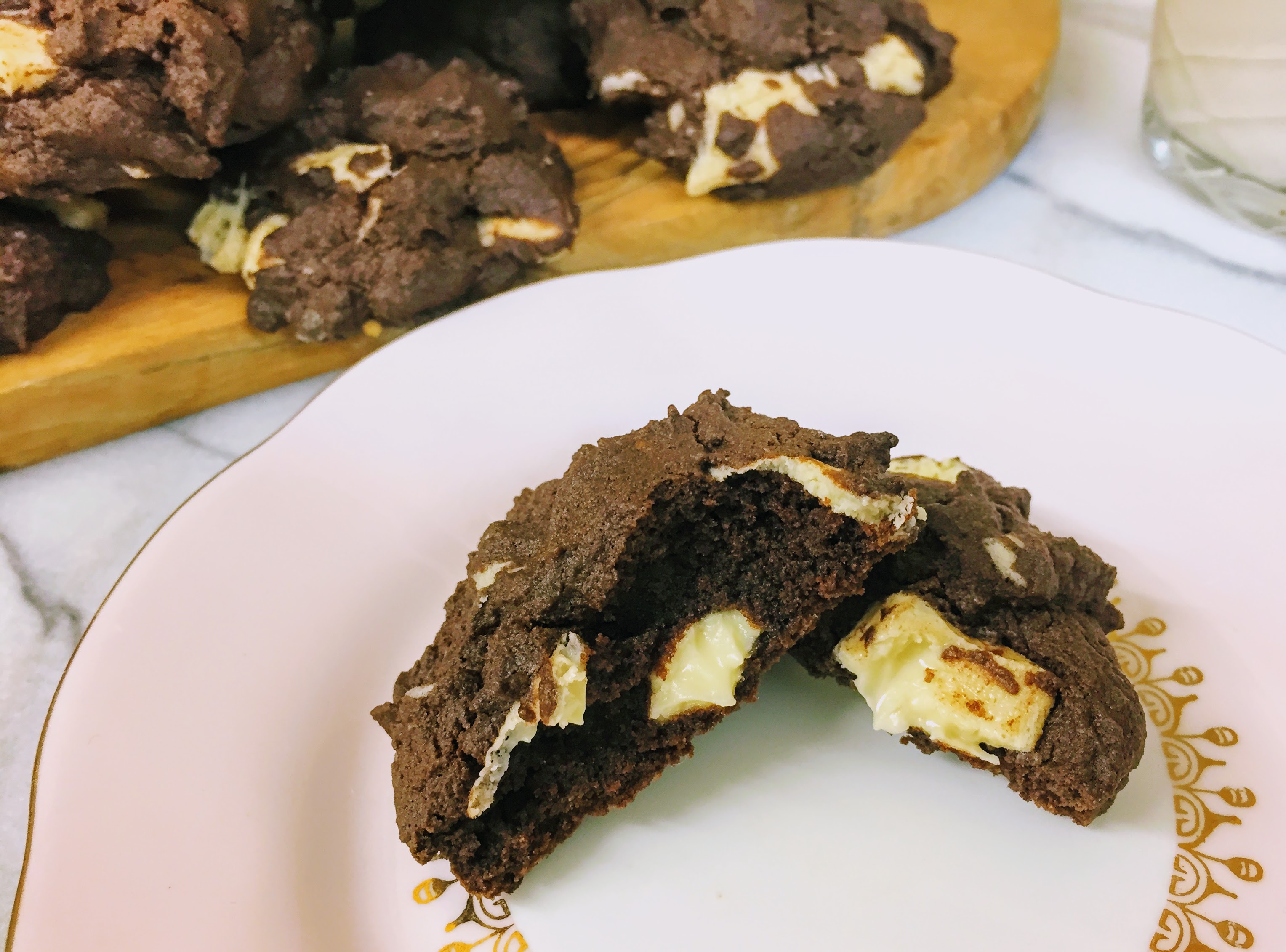 A double chocolate brownie cookie, split in half on a small plate, with a pile of cookies in the background. The outside of the cookie is crisp, but the inside is moist and fudgy, and there's a chunk of vegan white chocolate oozing at its center.