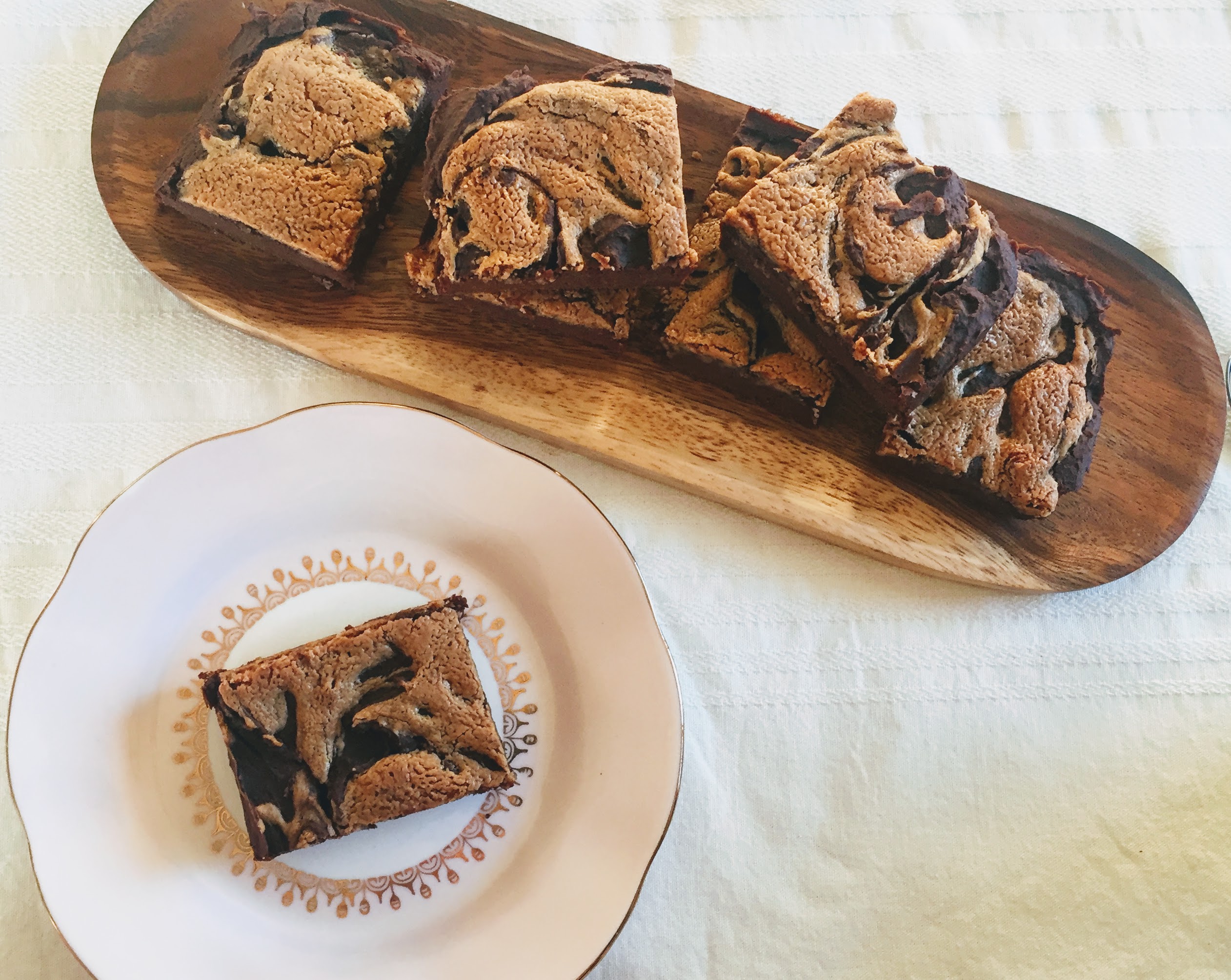 An aerial view of the peanut butter, banana and chickpea brownies. One is on a plate and the others are on a wooden board. You can see the golden peanut butter swirl topping contrasted with the deep, dark chocolate of the brownies.