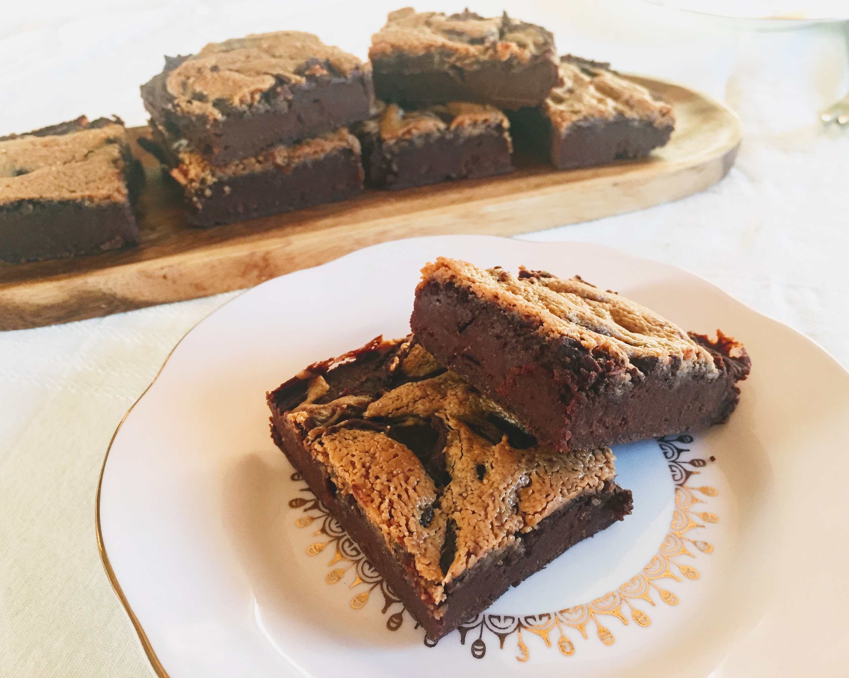 Two dark and fudgy brownies stacked on a pink place. Each has golden-coloured swirls of peanut butter on top, and the remaining brownies are on a platter in the background.
