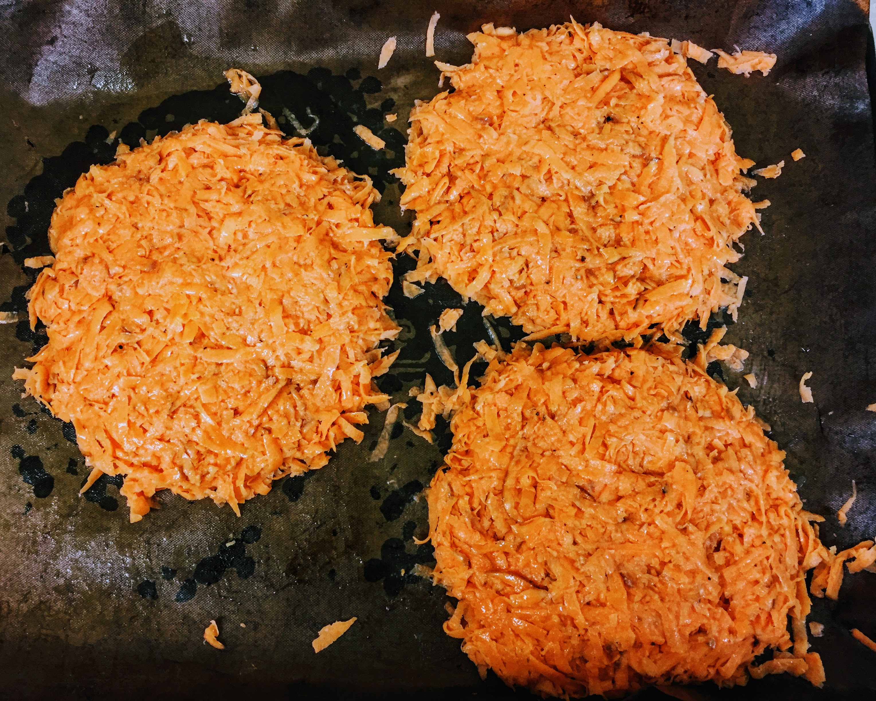 The bright orange grated sweet potato, pressed together to form three patties on a baking tray. The patties are roughly circular and level.
