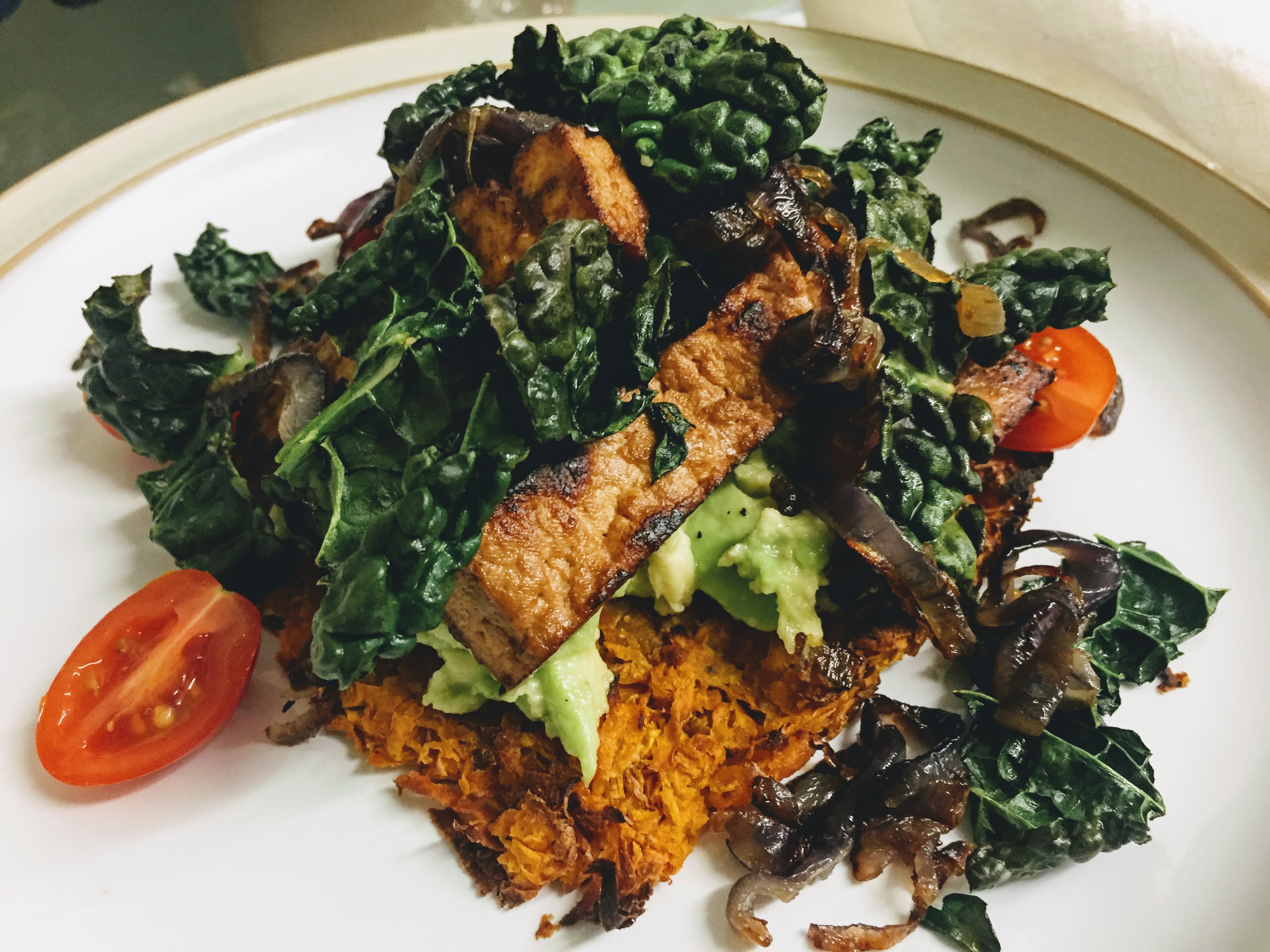 A side view of the finished dish. The sweet potato rösti is at the base, it's edges blackened slightly, and smothered in creamy smashed avocado. The fried tofu is nestled on top and everything is scattered with crispy kale and fried onions, with some cherry tomatoes peaking out.