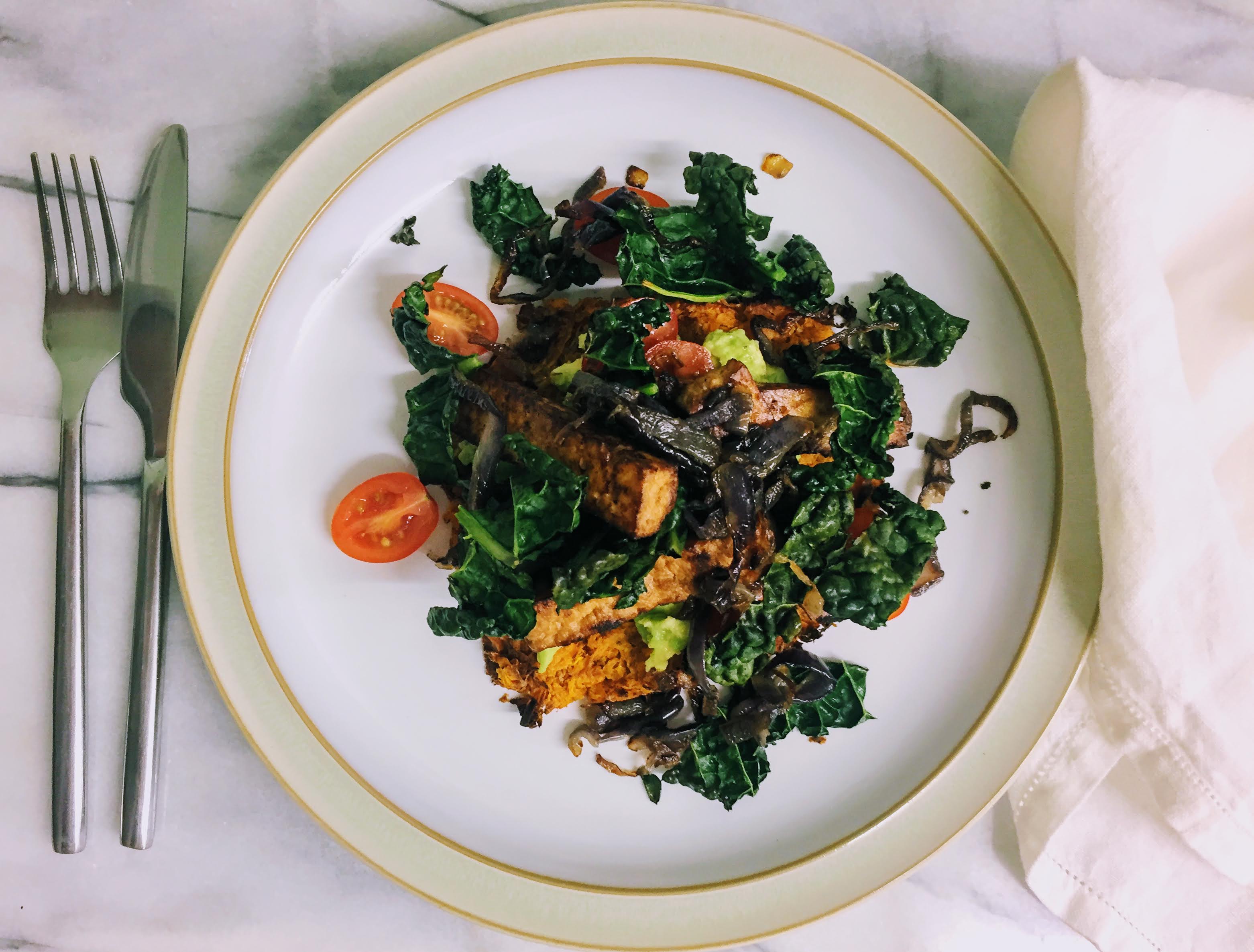 A white plate, photographed from above, with a tangle of dark green kale, caramelized red onions, cherry tomatotes, and charred batons of smoked tofu. There are flashes of lime green smashed avocado and the vibrant orange of the sweet potato rosti peaking out from underneath