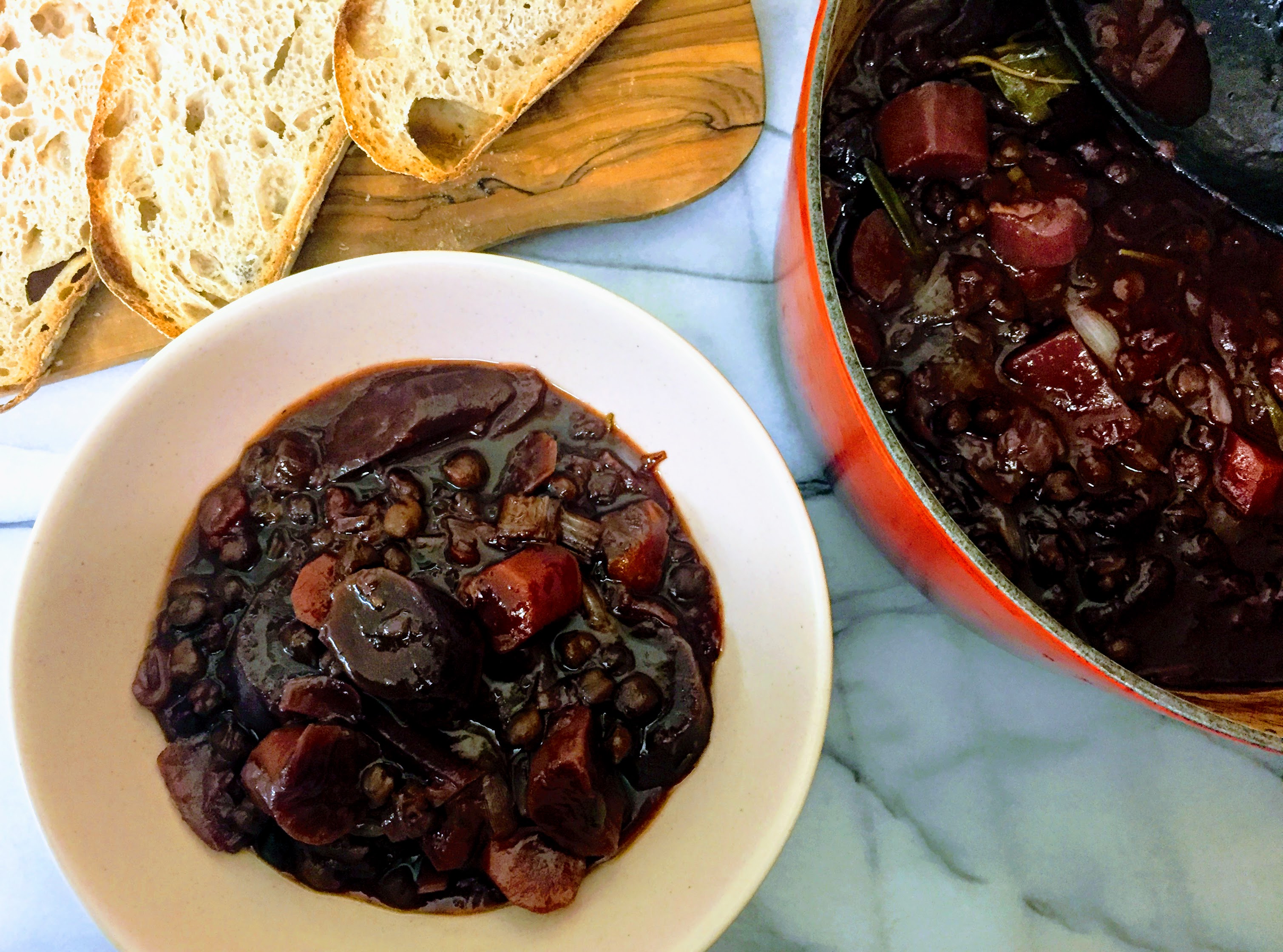 A bowl of chickpeas au vin seen from above. The stew has an intensely dark red wine colour, an there are large chunks of purple carrots surrounded by chickpeas, all drenched in smooth, dark and glossy gravy. To the right is the casserole dish the remaining stew, and at the top there are some thick slices of sourdough bread on a wooden board.