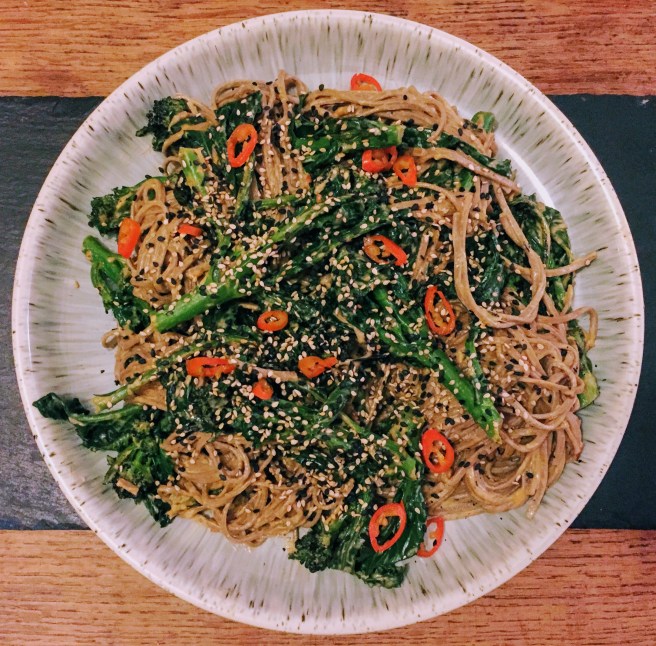 A large plate covered in a tangle of soba noodles and dark green spears of purple sprouting broccoli. Everything is coated in a creamy sesame sauce, and the entire thing is scattered with white and black toasted sesame seeds and bright flecks of red chilli. 