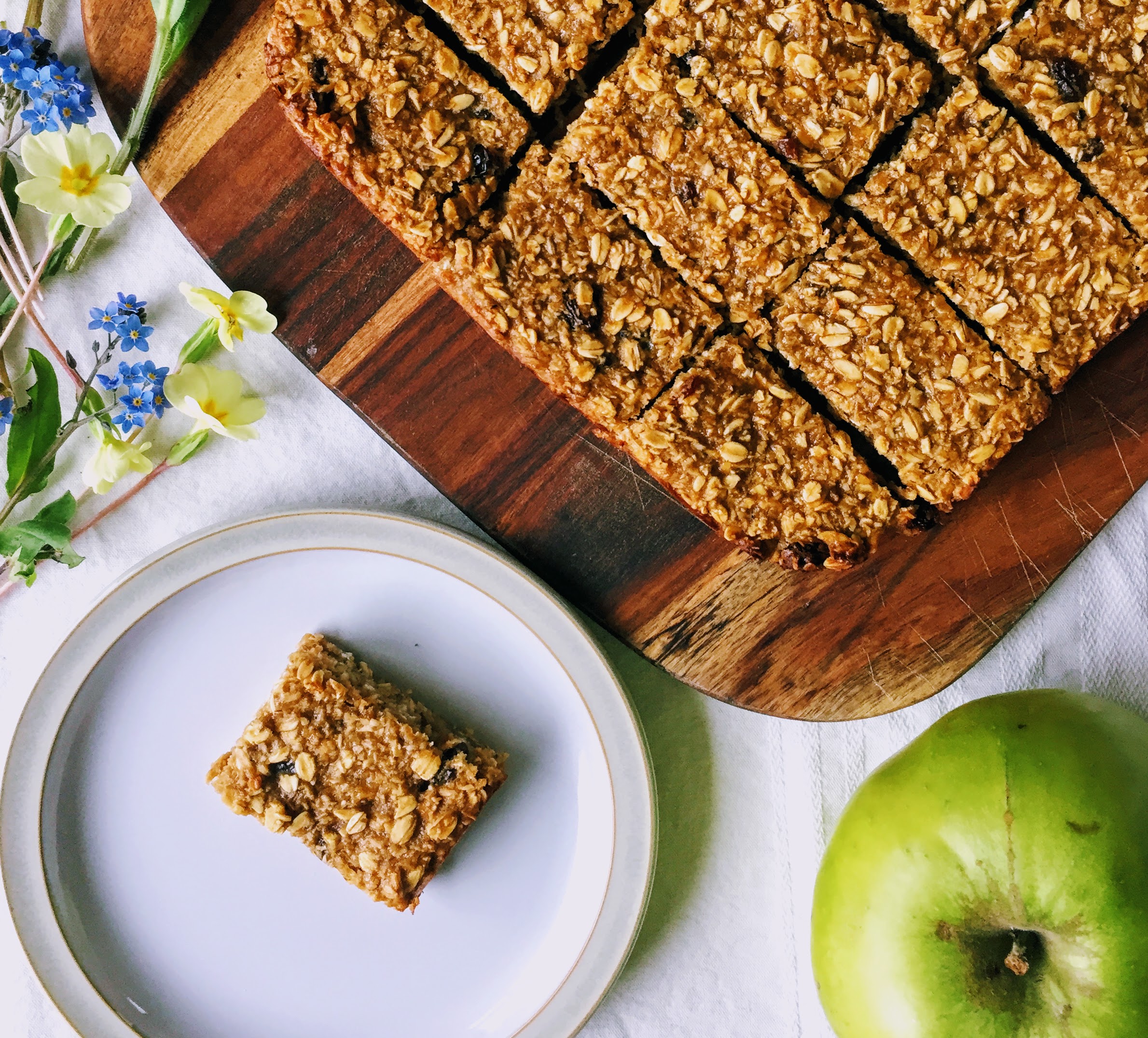 The flapjack slices, with one on a small plate at the bottom of the image. There's a large cooking apple at the bottom right, and some primroses and fort-get-me-nots to the left.