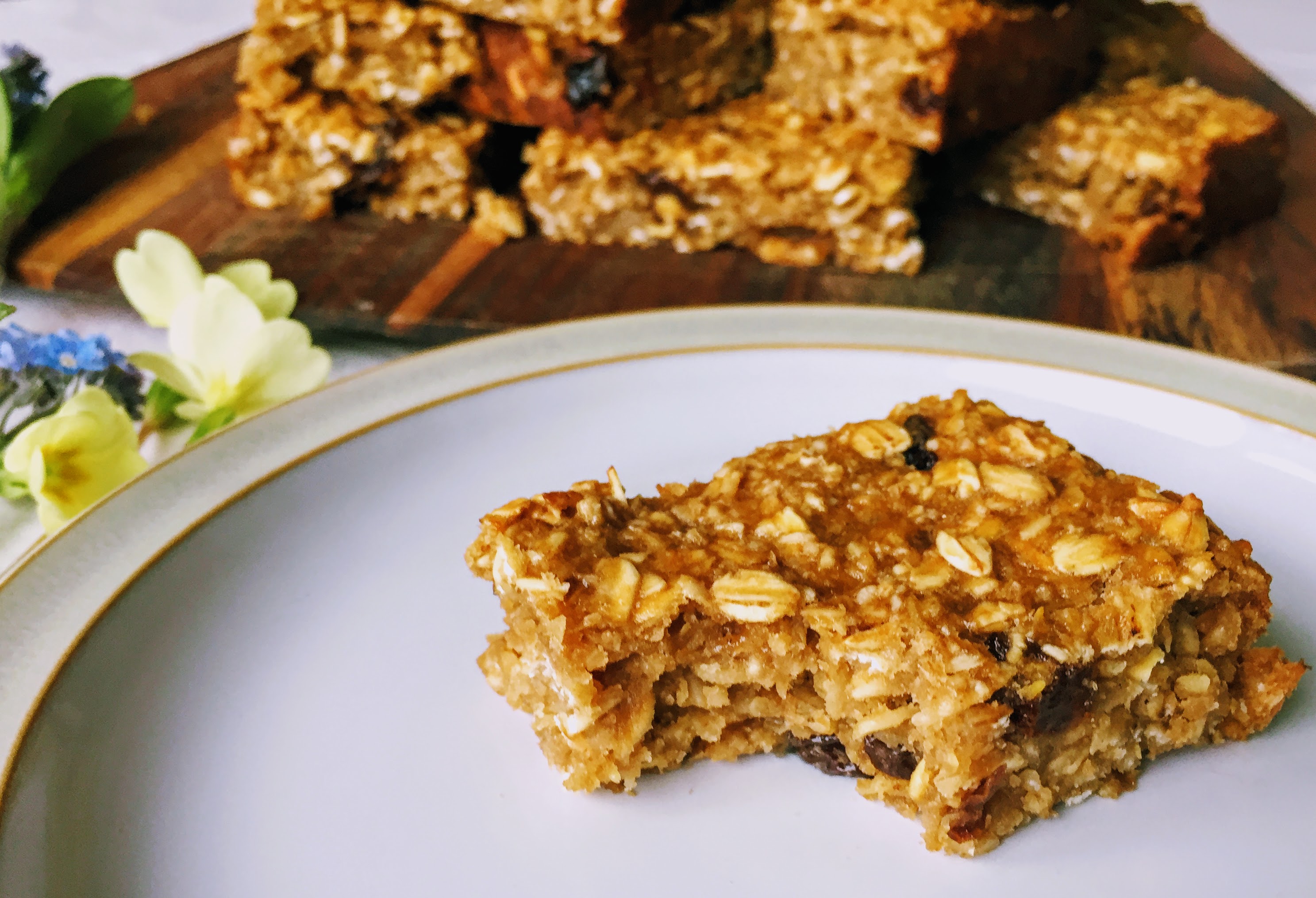 A close-up on a flapjack on plate with a bite taken out, to reveal a chewey, oaty centre. The remaining flapjacks are in a pile in the background with some spring flowers.