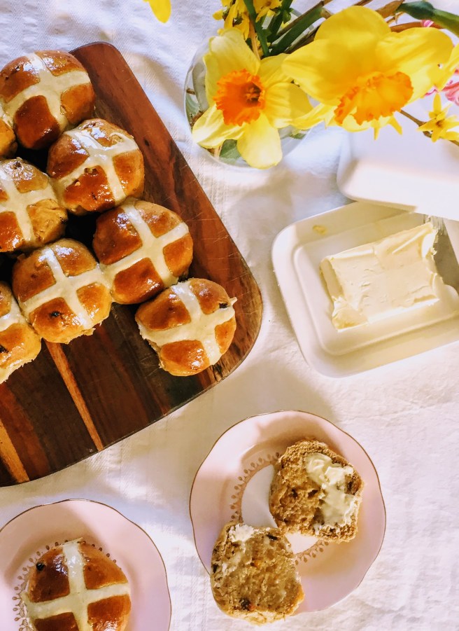 Shiny hot cross buns, served on a wooden board. There's a vase of bright yellow daffodils at the top of the picture, an open butter dish, and two hot cross buns on dainty pink plates, one of which has been cut in half and spread with lashings of butter. 