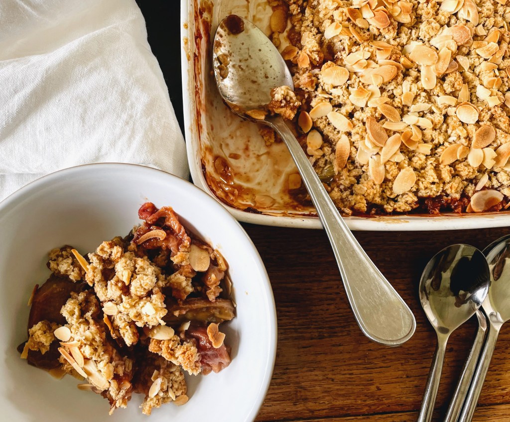 A serving of rhubarb crumble in a bowl, next to the remaining crumble in the oven dish. The pinks and greens of the rhubarb are peeking through and are beautifully soft, with chunks and clumps of golden oaty crumble on top.