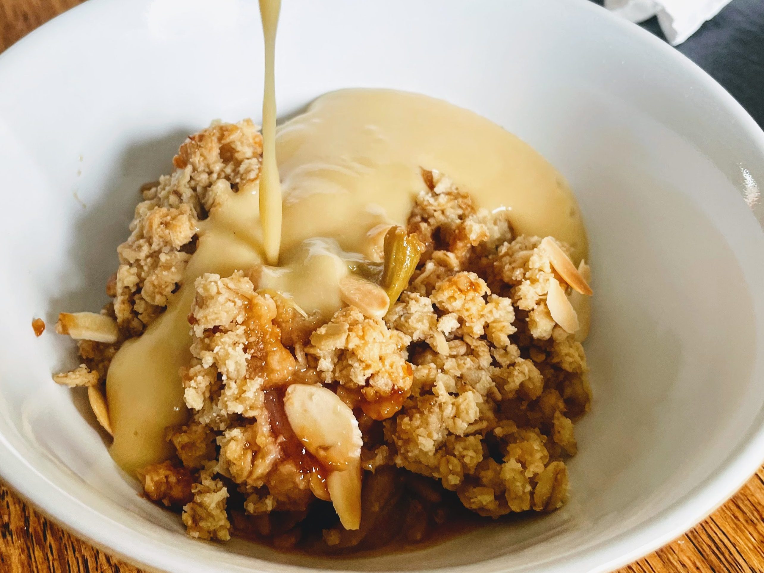 A bowl of rhubarb, orange and almond crumble, with smooth, thick vanilla custard being poured over the top.