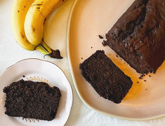 An aerial view of the sliced loaf of chocolate buckwheat banana bread. One slice remains on the plate next to the loaf, and the other is on a side plate nearby. There are a few dark chocolate crumbs and two bananas at the top left of the picture.
