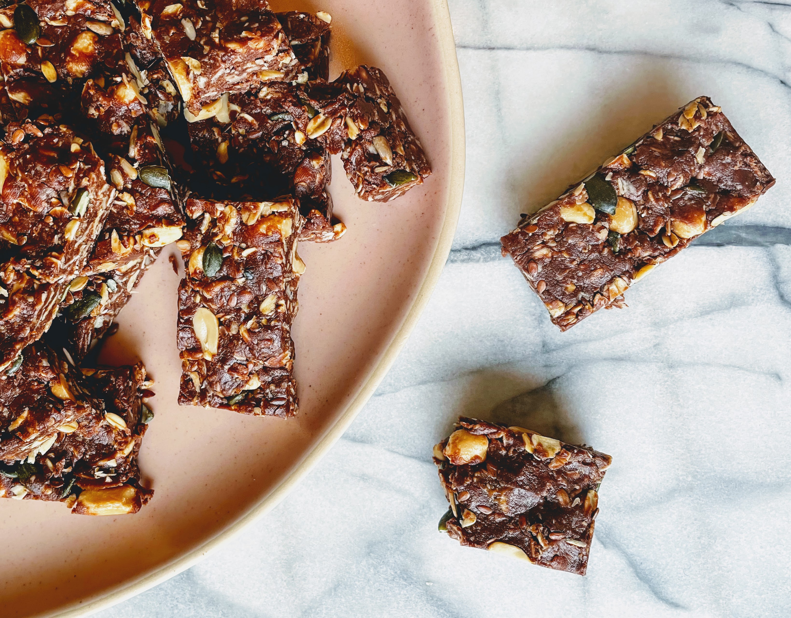 The chocolate and peanut bars on a pink plate, with a couple of the marble worktop besides. The bars are bursting with peanuts, seeds and oats, with a chocolate mixture holding them together. 