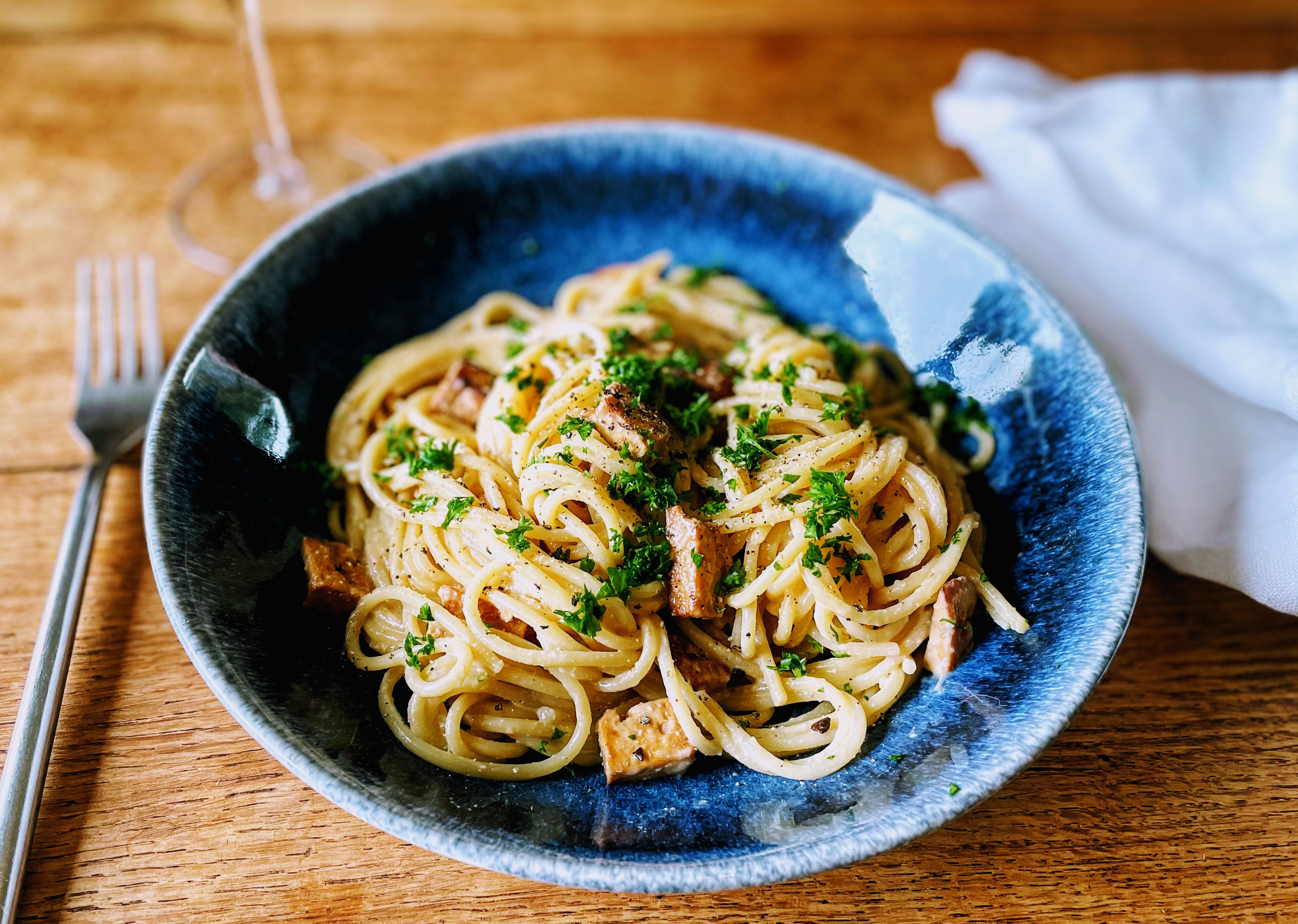 A dark blue shallow bowl piled with a tangle of creamy spaghetti, with little bits of crisp, marinated smoked tofu. There's a sprinkling of chopped fresh parsley on top.