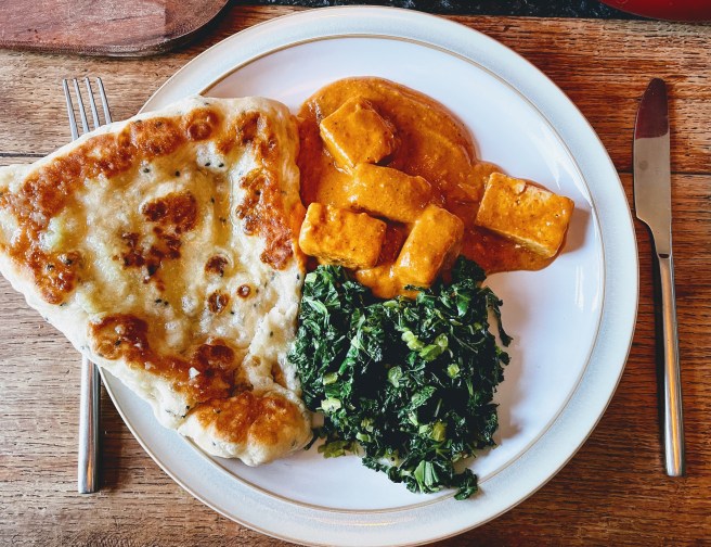 A plate of tofu butter masala, dark stir-fried kale and a fluffy naan with patches of deep golden brown. 