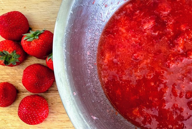 On the left hand side are some vibrant red strawberries scattered on a wooden board, and on the left is part of a metal bowl filled with crushed strawberries. You can still see some chunks of strawberry but the mixture is very wet, juicy and bright red.