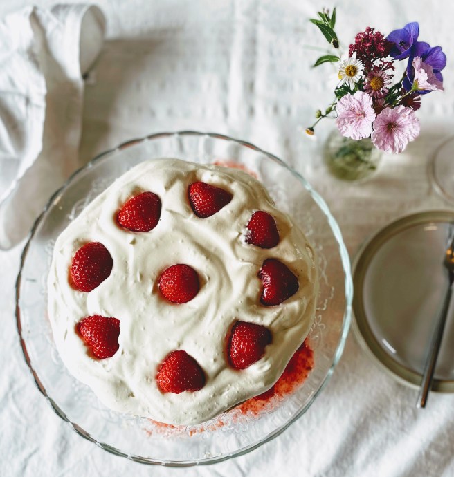 A view of the finished shortcake from above. The cream is softly swirled, and deep red strawberry halves are arranged in a circle around the edge, with one in the middle. The shortcake is on a class cake stand, and to the right is a side plate with a fork, and a small vase filled with delicate pink and purple flowers.