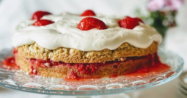 A closer, side-on view of the strawberry shortcake. The shortcake is light golden-brown and slightly rough in texture, and you can see that some of the juice from the crushed strawberry filling is starting to seep into the bottom layer of cake. The vegan whipped cream is piled on top, with a few jewel-red strawberry halves for decoration. 