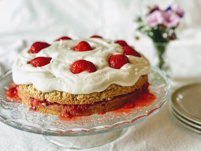 A strawberry shortcake on a glass cake stand.  The shortcake is pale golden brown, and sliced in half, with bright red crushed strawberries oozing out of the side. The top is slathered with soft and pillow-y whipped cream and dotted with bright and shiny strawberry halves. There are some side plates next to the cake stand, and a vase with pink and purple flowers in the background. 