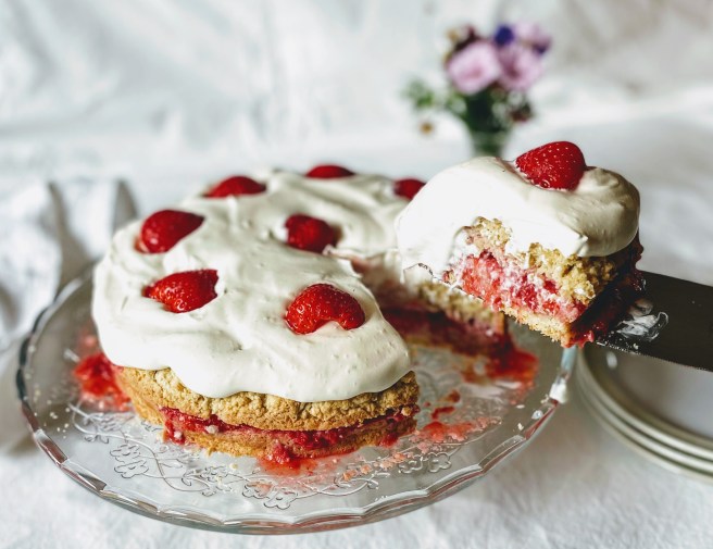 The shortcake with the first slice cut and balanced on a serving slice. You can see where the crushed strawberries have soaked into the bottom layer of cake, and the thick layer of soft cream has folded over the side. 