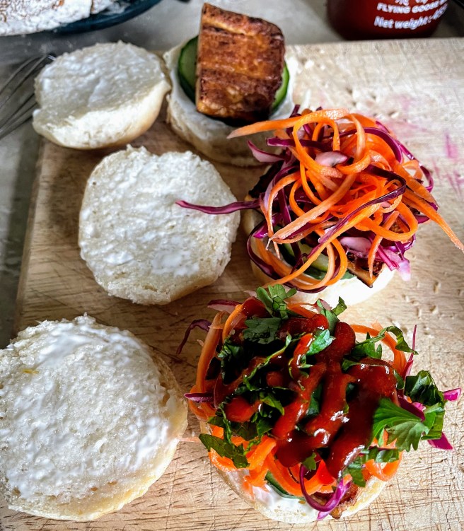 Three banh mi sliders being assembled. The bun furthest away from then camera is spread with mayonnaise and has a cucumber slice, then a piece of fried, smoked tofu. The middle bun has a generous tangle of pickle on top of the tofu, and the bun nearest the camera is ready for it's lid, with fresh herbs and a squirt of sriracha.