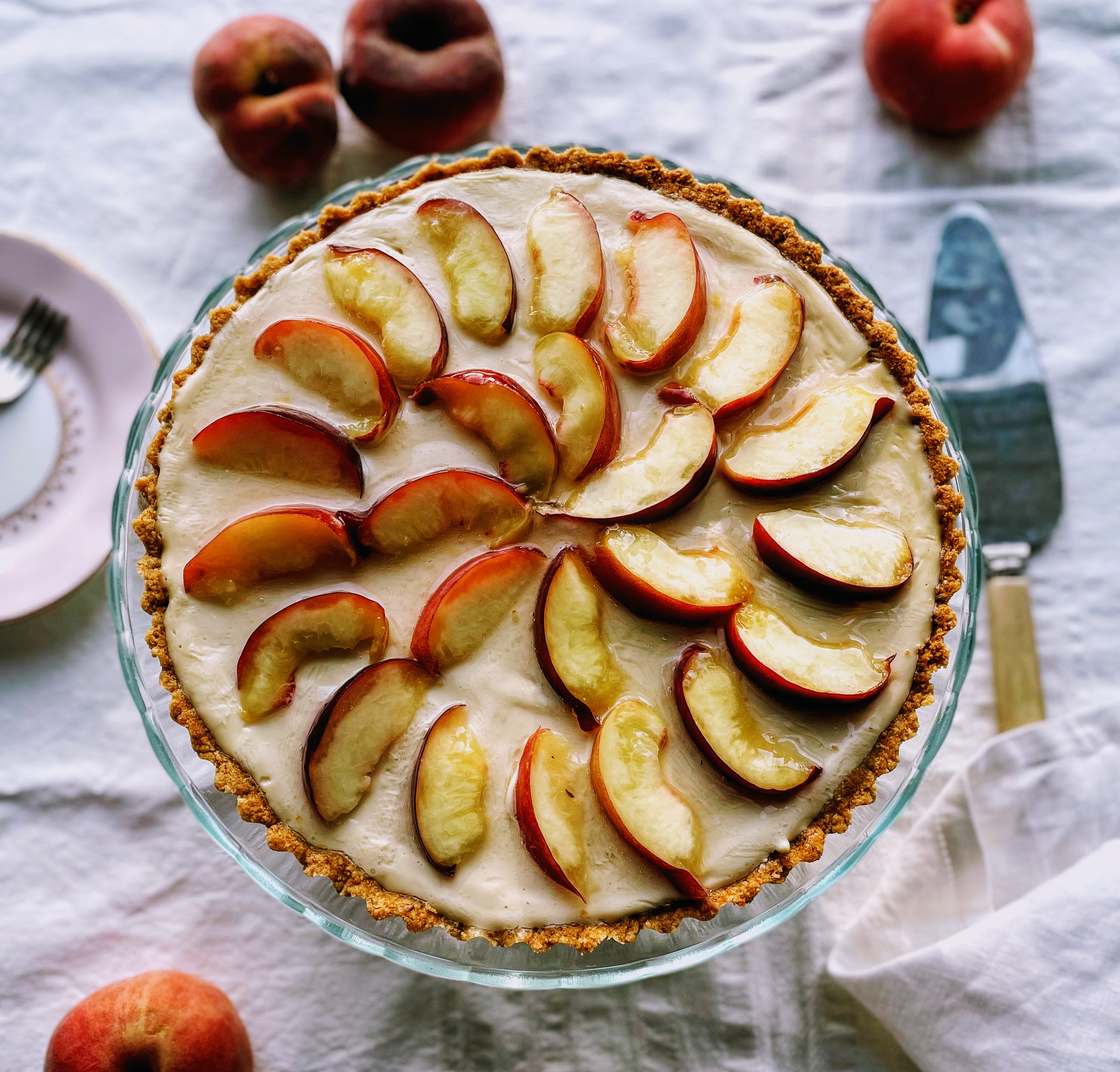 The complete peach and white chocolate tart on a cake stand. The edges are golden and creamy white chocolate filling is covered in fresh peach slices, arranged in a circle around the edge, and another circle in the middle. The top of the tart is shiny and glistening from the glaze.