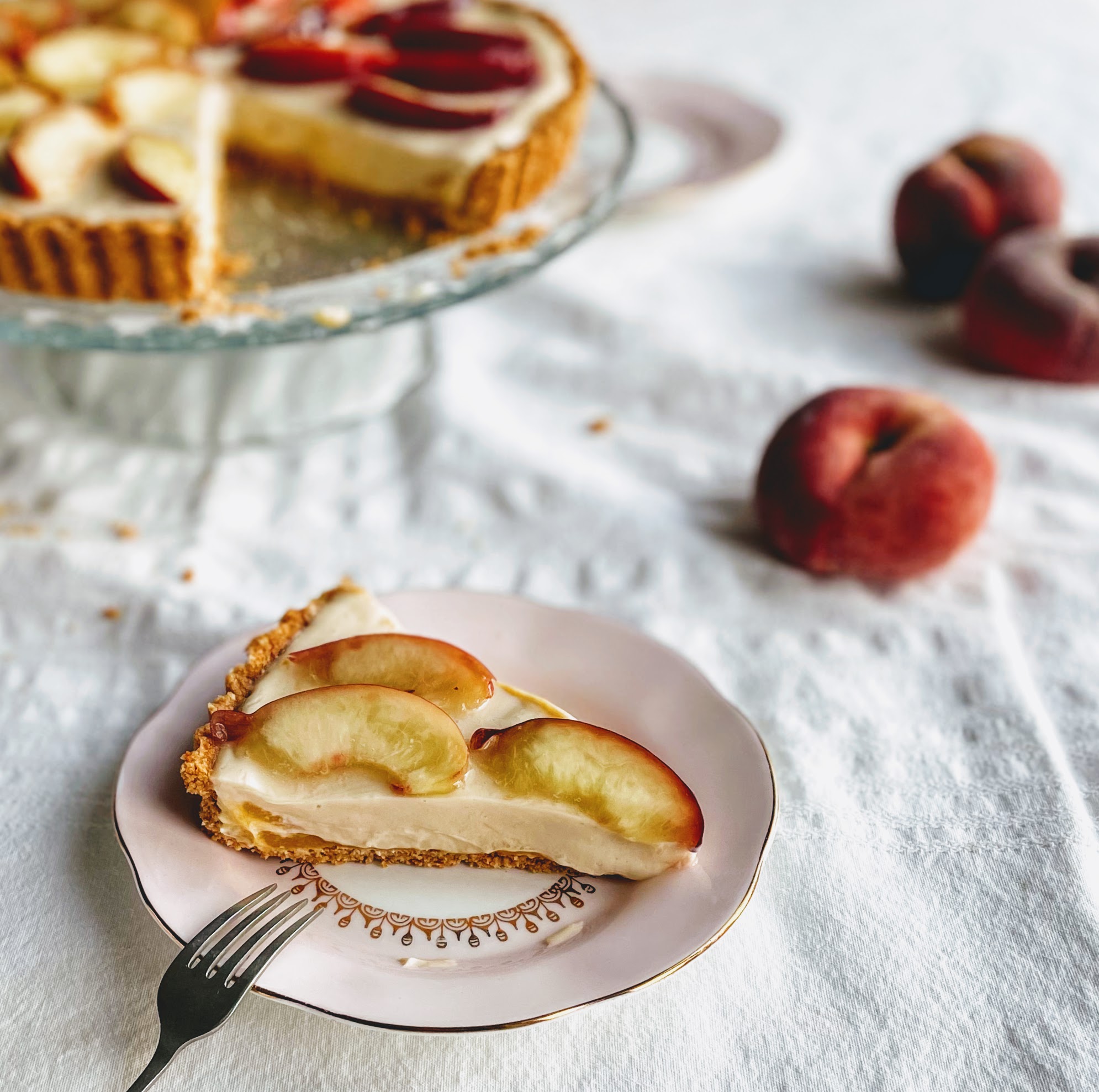 A slice of peach and white chocolate tart. The base is thin and crisp and you can see a bright flash of peach puree underneath the silky smooth white chocolate filling. There are three juicy slices of fresh peach on top, and three velvety peaches in the background, along with the rest of the tart on a glass cake stand. 