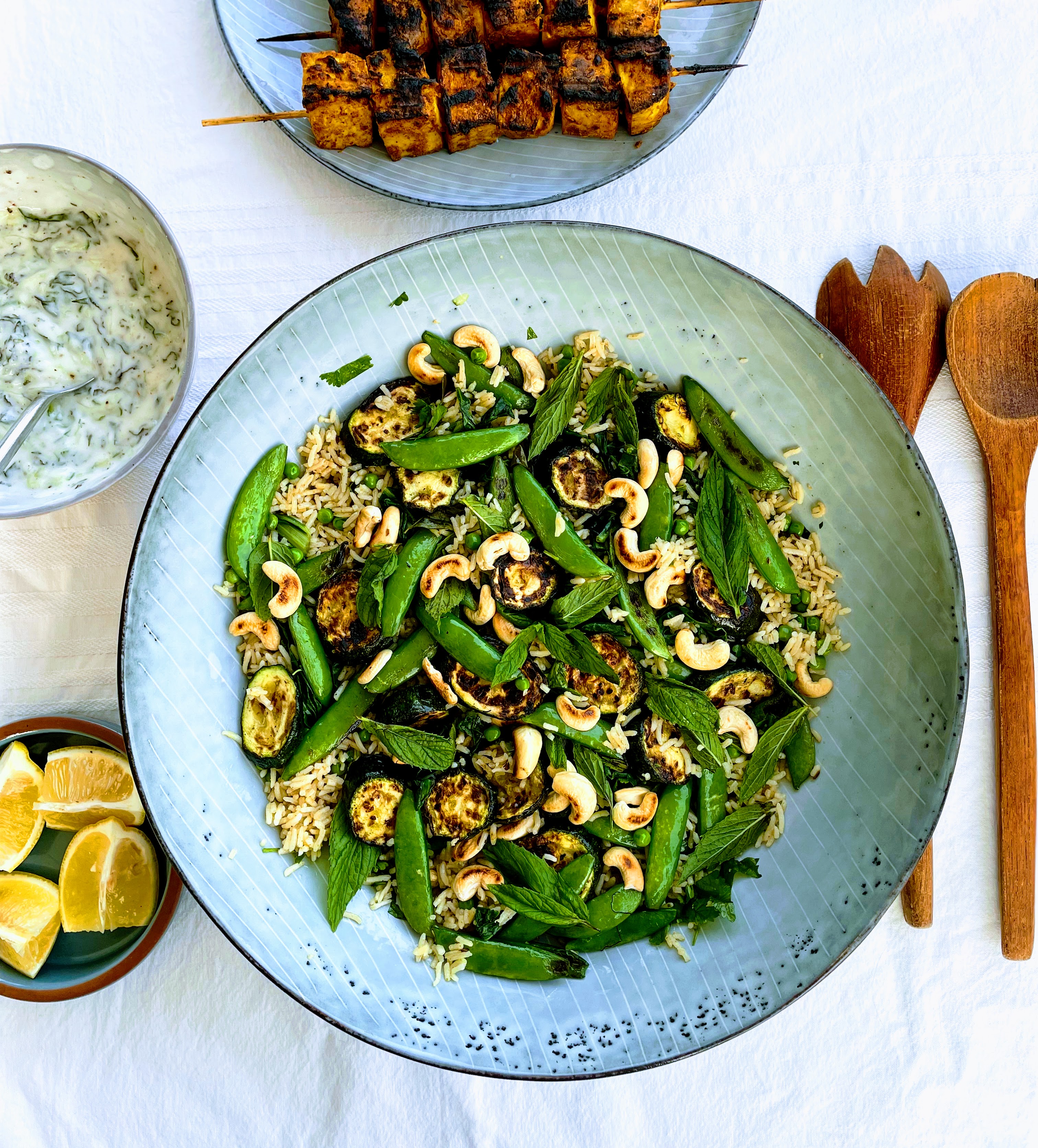 The serving bowl full of Indian rice salad seen from above. Vibrant green summer vegetables and herbs sit on top of delicately spiced rice and toasted cashews. There are lemon wedges in a bowl to the left, and wooden salad servers on the right. At the top is a bowl of creamy mint raita and some tandoori tofu skewers, blackened from the grill.  