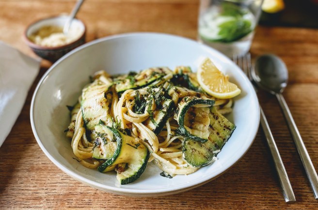 A white pasta bowl filled with a tangle of linguine and grilled slices of courgette. The courgette slices have scorch lines from the grill, and are soft, but still with a little rigidity. The pasta has a creamy sauce that clings to it, and is scattered with shredded mint leaves. There's a wedge of lemon on the side.