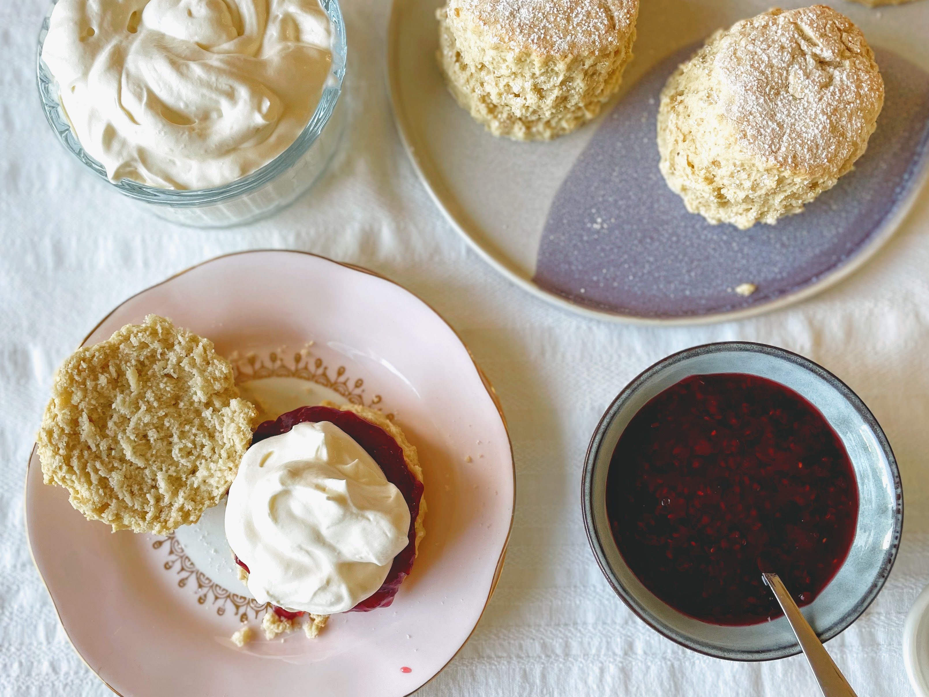 In the bottom left-hand corner of the picture there is a pale pink plate with a scone that has been split in two. One half reveals the soft, light crumb of the scone, and the other is smothered with crimson red raspberry jam and piled with whipped cream. There is a small bowl full of raspberry jam in the bottom right hand corner, and a dish of pillowy whipped cream in the top left. In the top right there is a blue patterned platter with the remaining scones, toweringly tall and with a little dusting of icing sugar on top. 