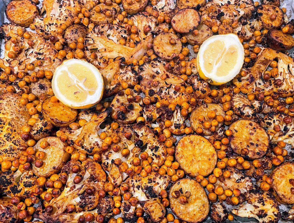 A large baking tray covered in a layer of roasted cauliflower, potatoes and chickpeas, with two lemon halves. The potatoes have turned golden, the chickpeas are crisp, and the edges of the cauliflower slices have blackened slightly. 