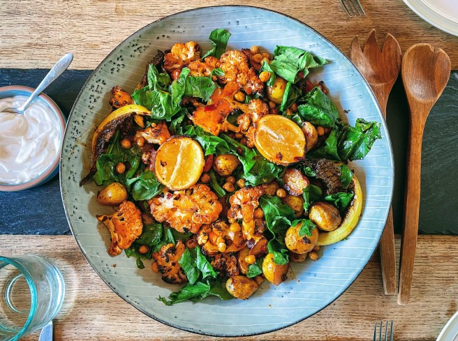 A large blue platter is seen from above, and filled with cauliflower, potatoes, and chickpeas, stained red from the harissa marinade, and tangled with bright green leaves of wilted spinach. There are two roasted lemon halves on top of the vegetables and a small dish of yogurt to the side. 