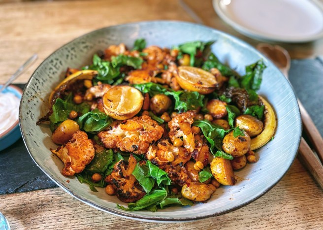 The blue platter of cauliflower and chickpea traybake from a side angle. The vegetables are all beautifully charred with the exception of the vibrant green wilted spinach.  
