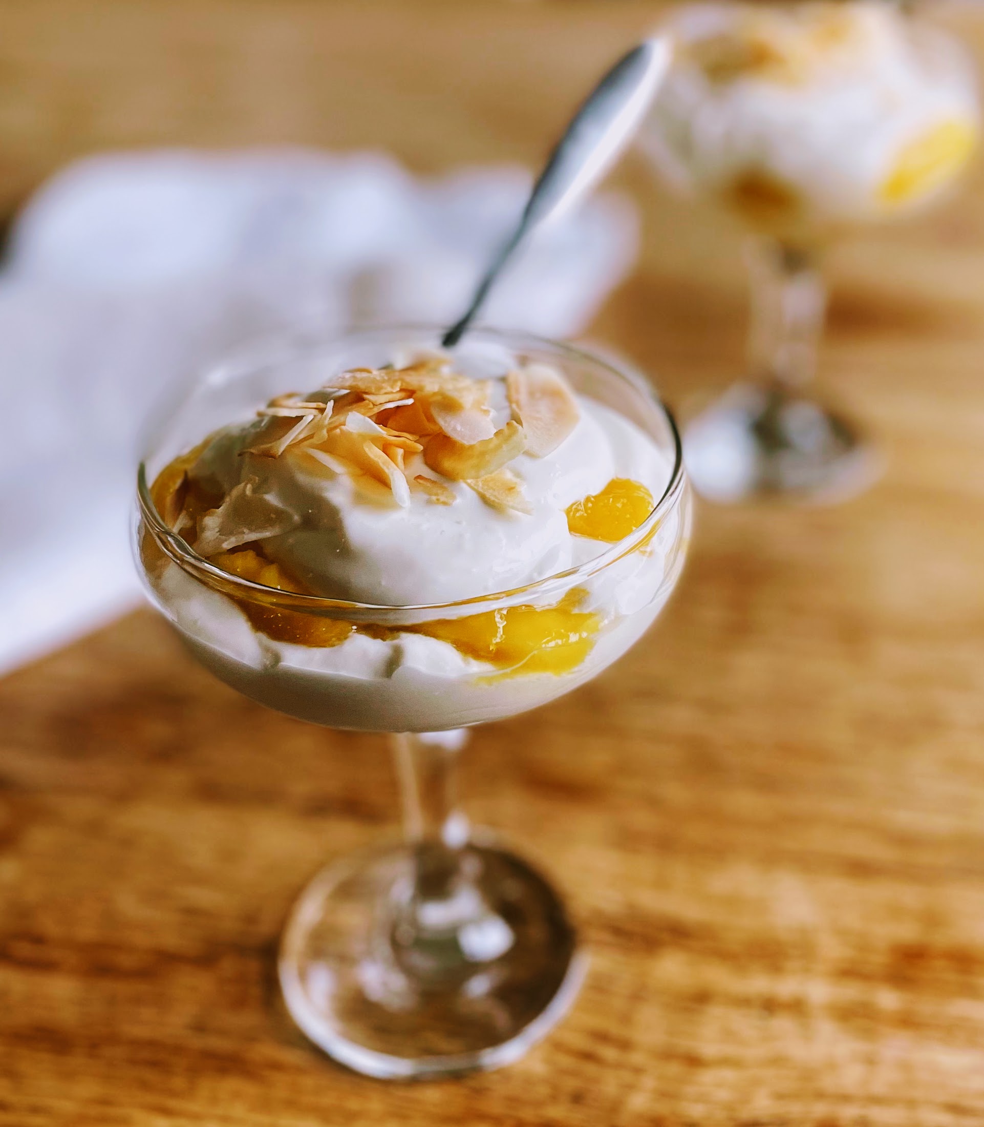 A glass coupe filled with mango and coconut syllabub. Vibrant golden mango slices peak out from under a pile of pillowy whipped yogurt and cream, with a scattering of toasted coconut flakes on top.